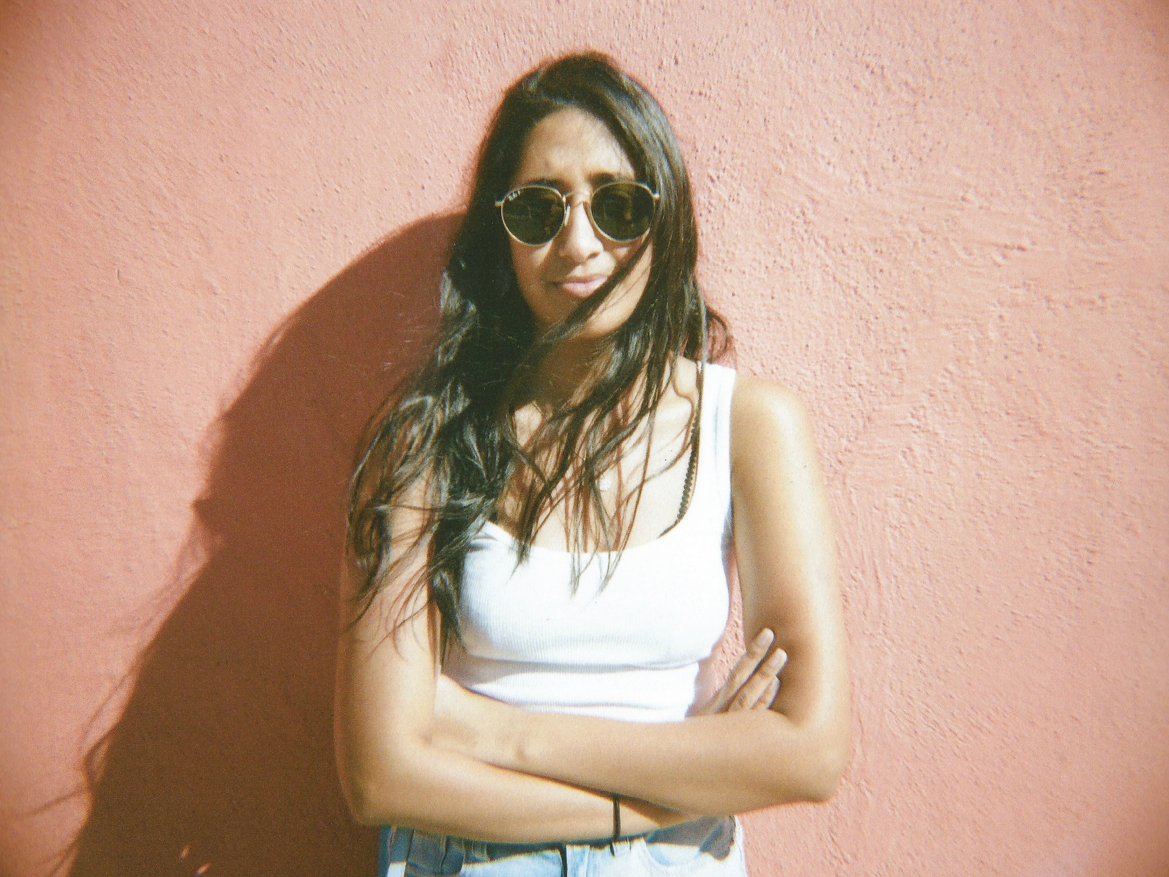 Portrait of Priya Parikh standing against a weathered terracotta wall, arms crossed, wearing round sunglasses, a white tank top, and denim, with long dark hair falling loose over one shoulder.